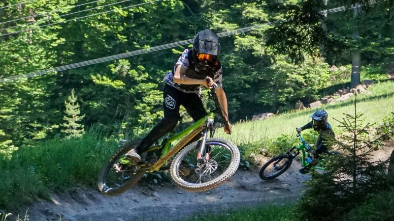 Zwei Mountainbiker mit Helmen fahren auf einem Waldweg im Adventure Bikepark Bad Wildbad. Einer springt, während der andere folgt., © Adventure Sports GmbH