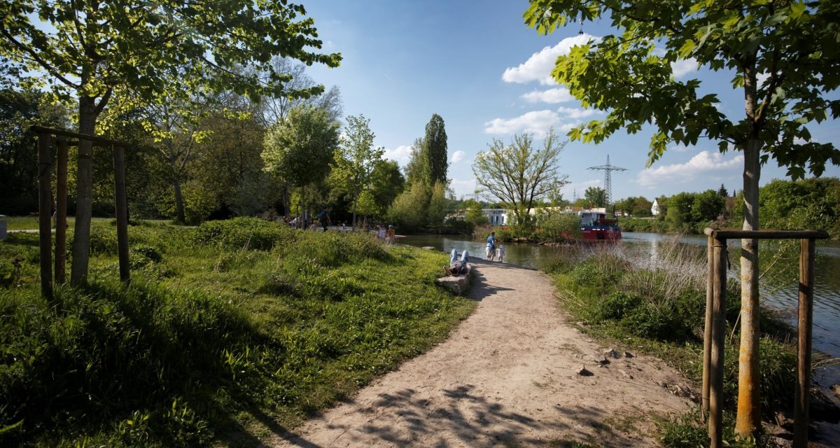 Ein sonniger Weg am Flussufer mit Bäumen und Spaziergängern. Der Himmel ist blau mit Wolken., © Stoppel Ein sonniger Weg am Flussufer mit Bäumen und Spaziergängern. Der Himmel ist blau mit Wolken., © Stoppel