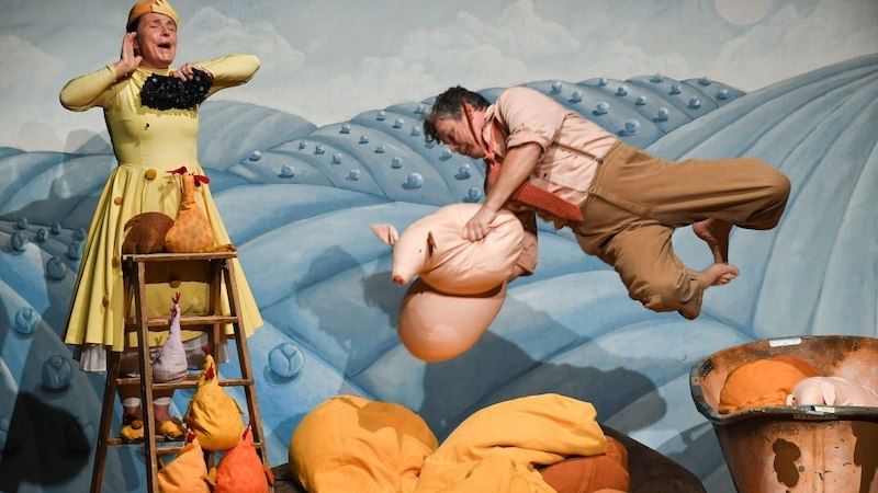 Bühnenbild mit zwei Schauspielern: Einer springt mit einem Stoffschwein, der andere steht auf einer Leiter. Bunte Hügel und Stofftiere im Hintergrund., © Stadt Kornwestheim Kulturmanagement