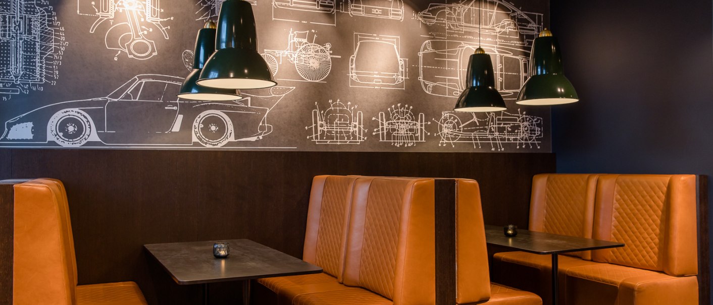 Restaurant mit braunen Ledersitzbänken, schwarzen Tischen und grünen Lampen. An der Wand sind technische Zeichnungen von Autos zu sehen., © TOMAS