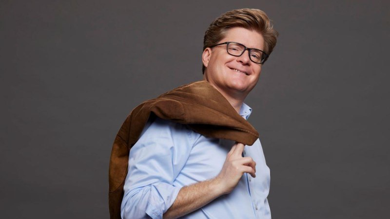 Ein Mann mit Brille trägt eine braune Jacke über der Schulter und lächelt. Er trägt ein hellblaues Hemd vor grauem Hintergrund., © Boris Breuer Ein Mann mit Brille trägt eine braune Jacke über der Schulter und lächelt. Er trägt ein hellblaues Hemd vor grauem Hintergrund., © Boris Breuer