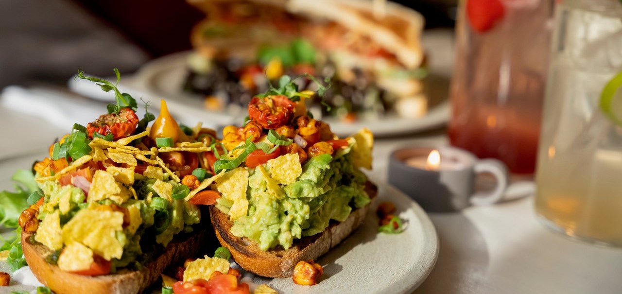 Avocado-Toast mit Tomaten und Kräutern, im Hintergrund ein Sandwich und Getränke. Ein Teelicht sorgt für gemütliche Atmosphäre., © Veganes Gold® Avocado-Toast mit Tomaten und Kräutern, im Hintergrund ein Sandwich und Getränke. Ein Teelicht sorgt für gemütliche Atmosphäre., © Veganes Gold®