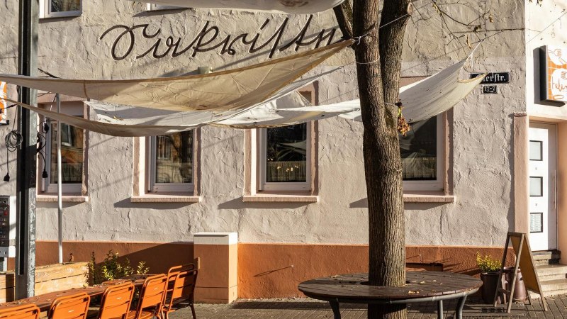 Ein Café mit Sonnensegeln und orangenen Stühlen im Außenbereich. Die Fassade ist beige mit Schriftzug. Ein Baum steht vor dem Eingang., © Stuttgart-Marketing GmbH, Sarah Schmid Ein Café mit Sonnensegeln und orangenen Stühlen im Außenbereich. Die Fassade ist beige mit Schriftzug. Ein Baum steht vor dem Eingang., © Stuttgart-Marketing GmbH, Sarah Schmid