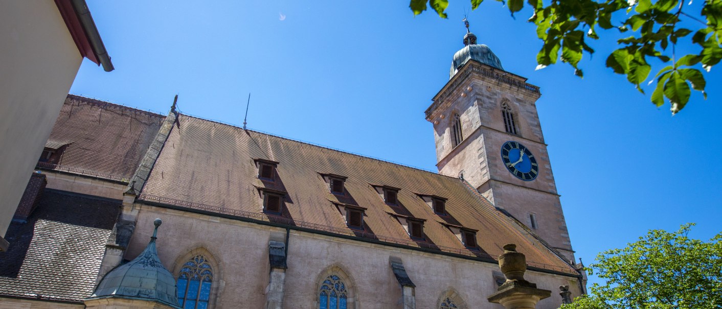 Die Nürtinger Stadtkirche St. Laurentius mit ihrem markanten Turm und einer großen Uhr, umgeben von Bäumen und strahlend blauem Himmel., © SMG, A. Mende