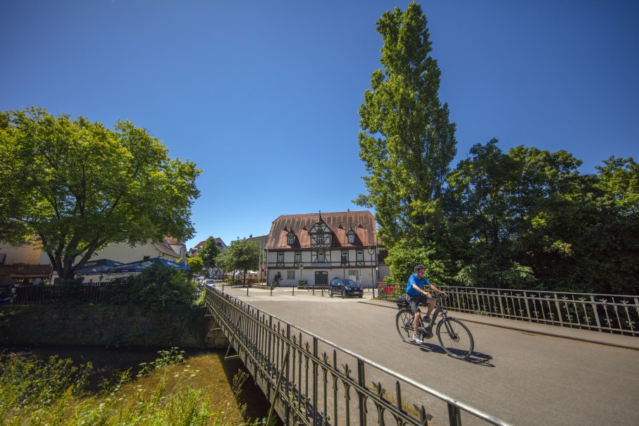Ein Radfahrer überquert eine Brücke in einer sonnigen Stadtlandschaft. Im Hintergrund steht ein Fachwerkhaus, umgeben von Bäumen., © SMG, A. Mende