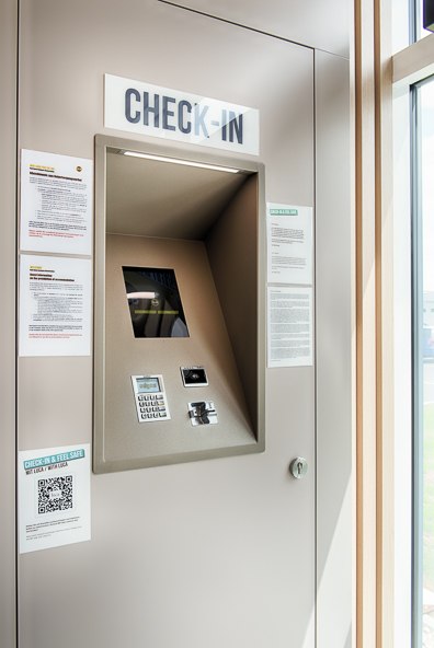 Ein moderner Check-in-Kiosk mit Bildschirm und Tastatur, umgeben von Informationsblättern und einem QR-Code, in einem hellen Raum., © B&B Hotel Stuttgart-Neckarhafen