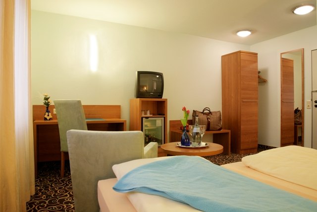 Gemütliches Hotelzimmer mit Doppelbett, Schreibtisch, Fernseher, Minibar und Tisch mit Blumen. Helle Wände und Teppichboden., © ateckhotel Gemütliches Hotelzimmer mit Doppelbett, Schreibtisch, Fernseher, Minibar und Tisch mit Blumen. Helle Wände und Teppichboden., © ateckhotel