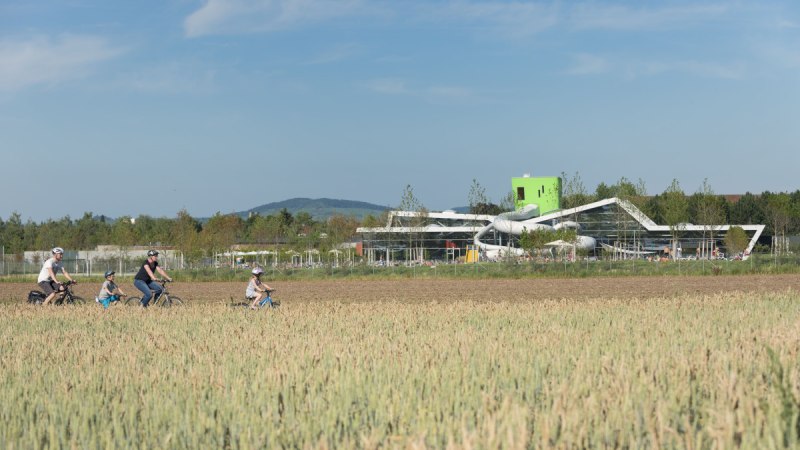 Familie radelt durch ein Feld, modernes Geb&auml;ude mit Rutschen im Hintergrund, blauer Himmel., &copy; Stadt Fellbach