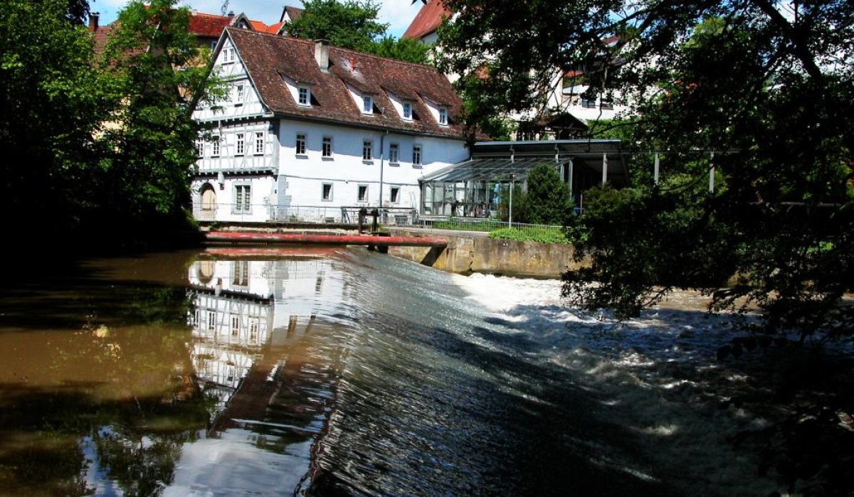 Historische Mühle mit Fachwerk und rotem Dach an einem Fluss mit Wehranlage, umgeben von Bäumen und blauem Himmel., © Waiblingen - Stuttgart-Marketing GmbH Historische Mühle mit Fachwerk und rotem Dach an einem Fluss mit Wehranlage, umgeben von Bäumen und blauem Himmel., © Waiblingen - Stuttgart-Marketing GmbH