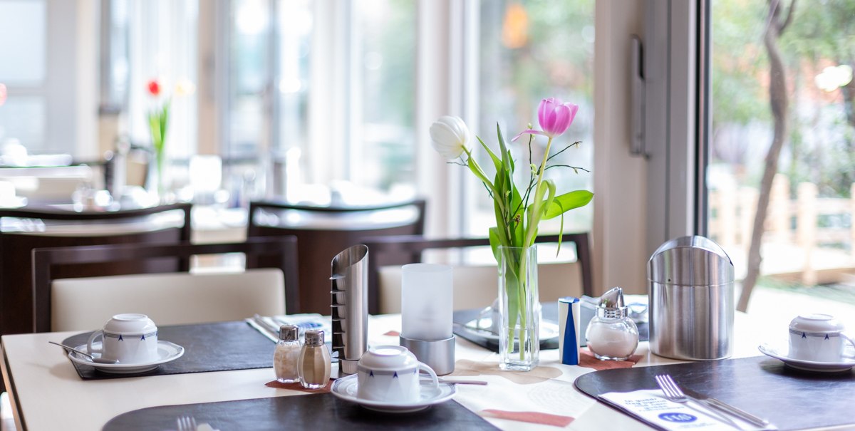 Ein Tisch in einem Café ist mit Geschirr, Besteck und einer Vase mit Tulpen gedeckt. Im Hintergrund sind Fenster und weitere Tische zu sehen., © TOMAS Ein Tisch in einem Café ist mit Geschirr, Besteck und einer Vase mit Tulpen gedeckt. Im Hintergrund sind Fenster und weitere Tische zu sehen., © TOMAS
