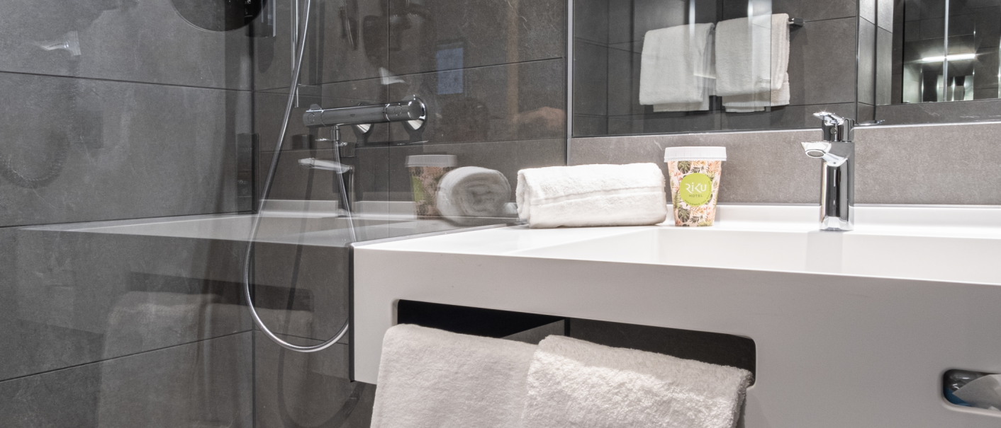 Modernes Badezimmer mit grauen Fliesen, einem weißen Waschbecken, Handtüchern und einem Becher mit Hotel-Logo auf der Ablage., © 13 6 Design GmbH (Judenhof 10, 89073 Ulm) Modernes Badezimmer mit grauen Fliesen, einem weißen Waschbecken, Handtüchern und einem Becher mit Hotel-Logo auf der Ablage., © 13 6 Design GmbH (Judenhof 10, 89073 Ulm)