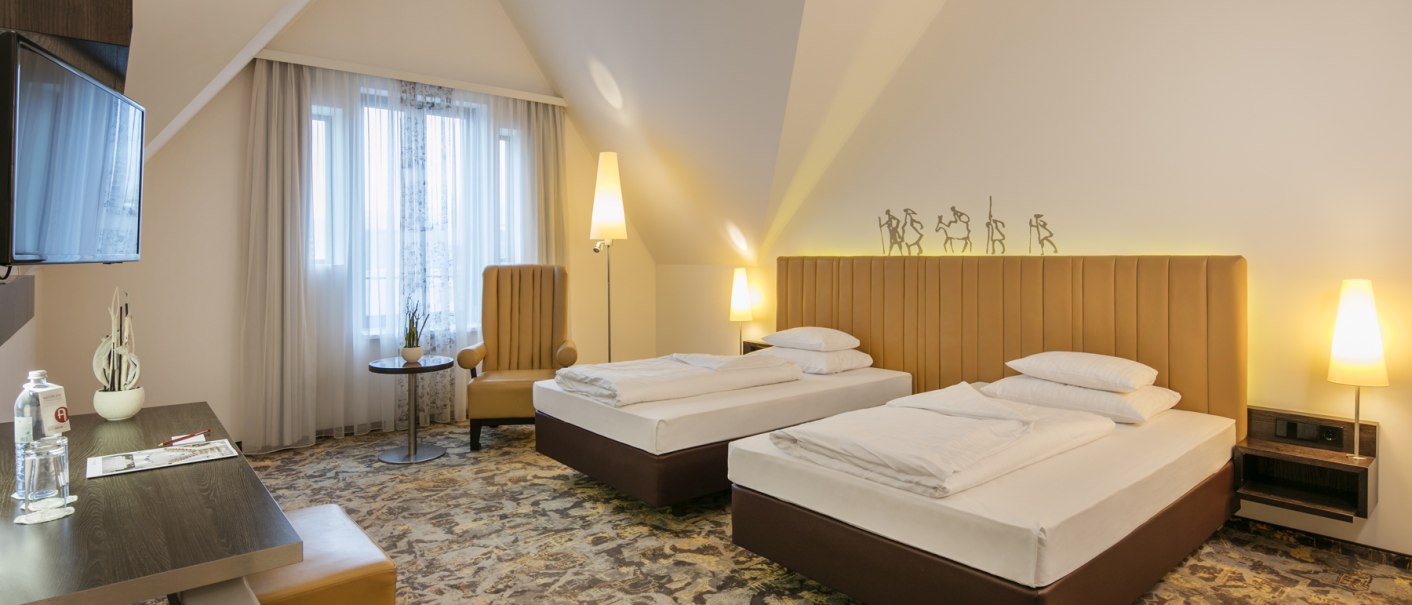 Ein modernes Hotelzimmer mit zwei Einzelbetten, einem Schreibtisch, einem Sessel und einem Flachbildfernseher. Die Einrichtung ist in warmen T&ouml;nen gehalten., &copy; ARCOTEL Hotels