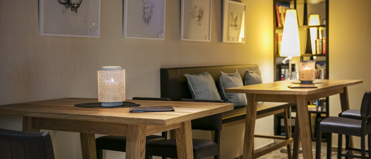 Gem&uuml;tliche Bar mit Holztischen, Kerzenlicht und Kunst an den W&auml;nden. Im Hintergrund ein B&uuml;cherregal und dezente Beleuchtung., &copy; Arcotel Hotels