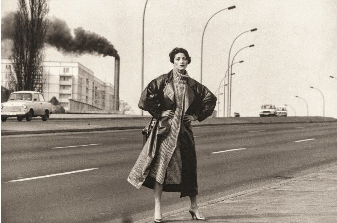 Eine Frau in einem langen Mantel posiert auf einer leeren Straße. Im Hintergrund sind rauchende Schornsteine und Autos zu sehen, was eine industrielle Umgebung andeutet., © Kunsthalle Göppingen Eine Frau in einem langen Mantel posiert auf einer leeren Straße. Im Hintergrund sind rauchende Schornsteine und Autos zu sehen, was eine industrielle Umgebung andeutet., © Kunsthalle Göppingen