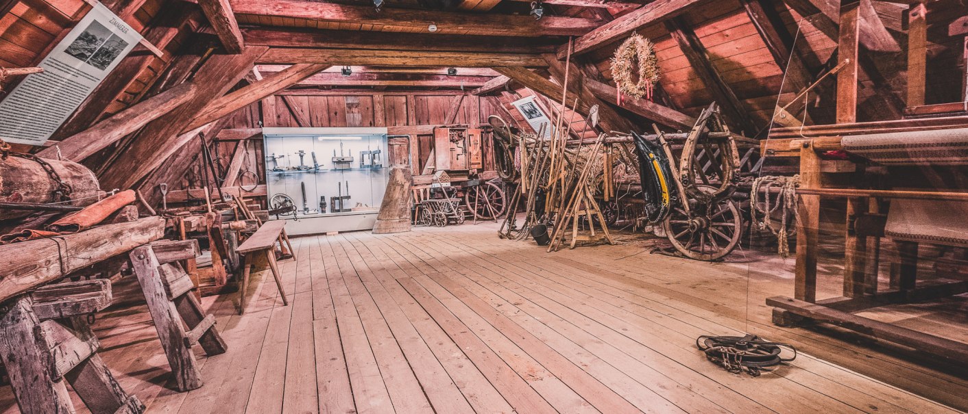 Ein rustikaler Dachboden mit Holzbalken, gefüllt mit alten Werkzeugen und landwirtschaftlichen Geräten. Ein Schaukasten zeigt weitere Exponate., © Stadt Welzheim Ein rustikaler Dachboden mit Holzbalken, gefüllt mit alten Werkzeugen und landwirtschaftlichen Geräten. Ein Schaukasten zeigt weitere Exponate., © Stadt Welzheim