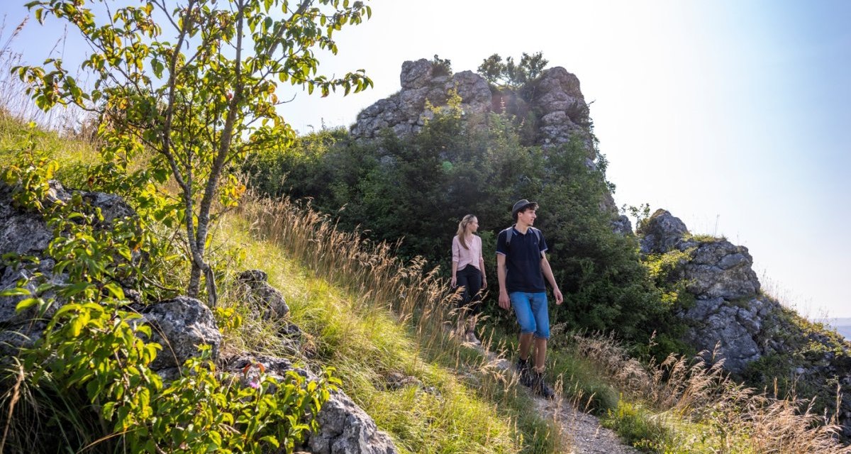 Zwei Personen wandern auf einem schmalen Pfad neben Felsen und Bäumen. Die Sonne scheint, und das Gras ist hoch gewachsen., © TMBW, Gregor Lengler Zwei Personen wandern auf einem schmalen Pfad neben Felsen und Bäumen. Die Sonne scheint, und das Gras ist hoch gewachsen., © TMBW, Gregor Lengler