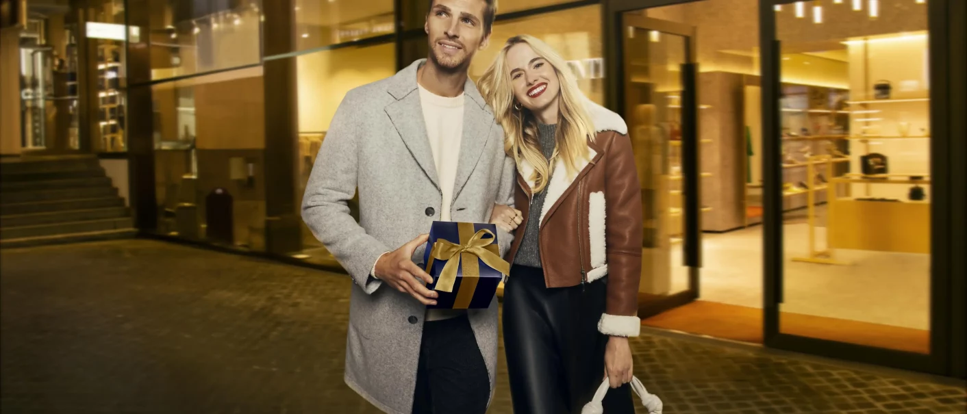 L&auml;chelndes Paar vor einem beleuchteten Gesch&auml;ft, der Mann mit Geschenkbox, die Frau mit Handtasche. Weihnachtlich und elegant., &copy; OUTLETCITY AG