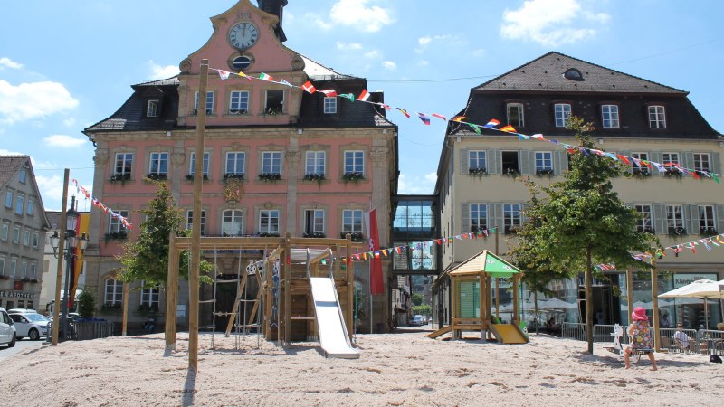 Ein Spielplatz mit Rutsche und Kletterger&uuml;st auf einem sandigen Platz vor historischen Geb&auml;uden, geschm&uuml;ckt mit bunten Wimpeln unter blauem Himmel., &copy; F&uuml;r die Bildrechte: Amt f&uuml;r Medien und Kommunikation Schw&auml;bisch Gm&uuml;nd