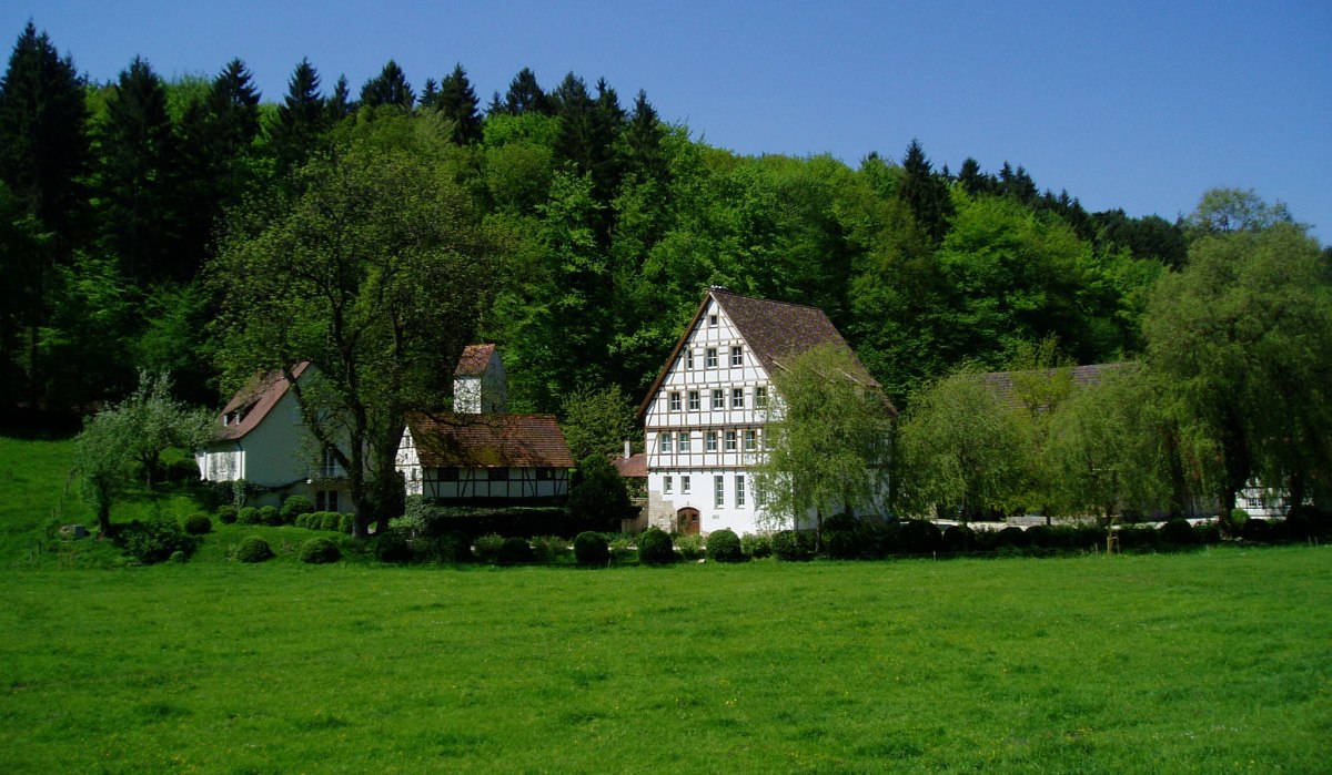 Fachwerkhäuser stehen in einer grünen Wiese vor einem dichten Wald. Der Himmel ist klar und blau., © Natur.Nah. Schönbuch & Heckengäu Fachwerkhäuser stehen in einer grünen Wiese vor einem dichten Wald. Der Himmel ist klar und blau., © Natur.Nah. Schönbuch & Heckengäu