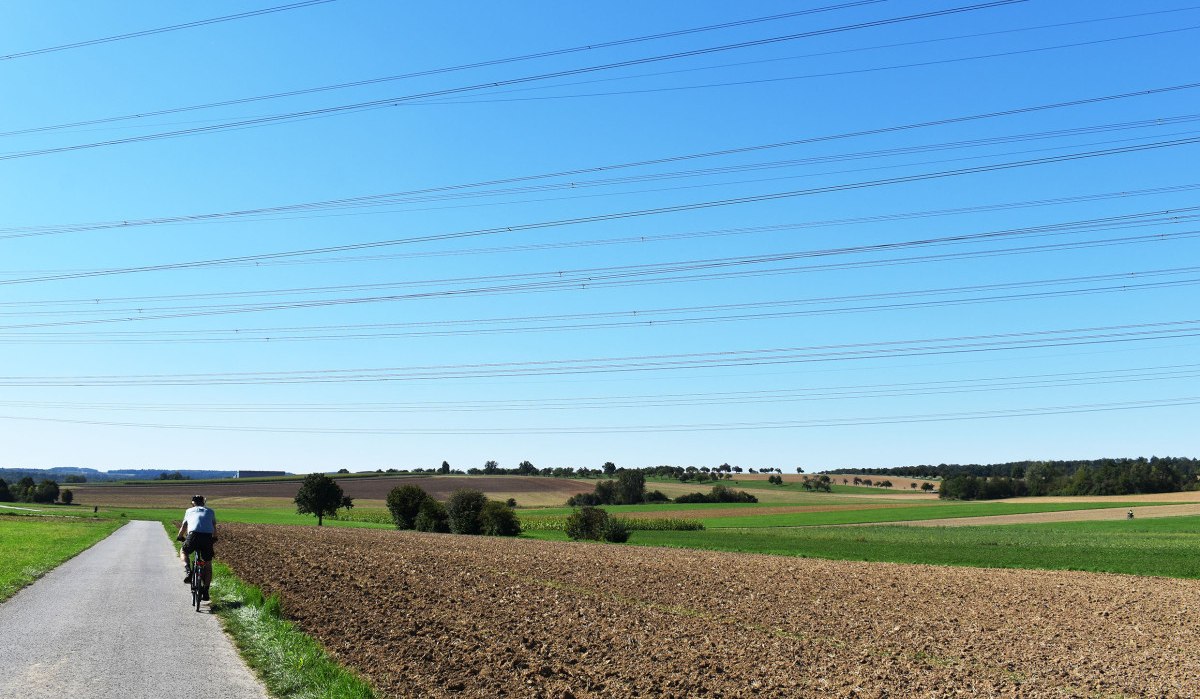 Ein Radfahrer fährt auf einem ländlichen Weg entlang von Feldern. Der Himmel ist klar und blau, darüber verlaufen Stromleitungen., © Stadtmarketing Böblingen e.V. Ein Radfahrer fährt auf einem ländlichen Weg entlang von Feldern. Der Himmel ist klar und blau, darüber verlaufen Stromleitungen., © Stadtmarketing Böblingen e.V.