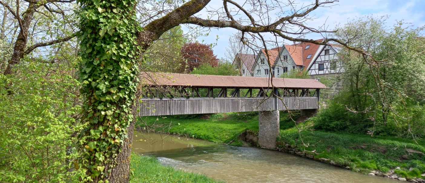 Fluss mit überdachter Holzbrücke, am Ufer Büsche und Bäume, auf der rechten Bildseite sind die weißen Giebelseiten und rotbraunen Dachflächen von vier Häusern hinter der Holzbrücke zu erkennen, © Stadt Gaildorf