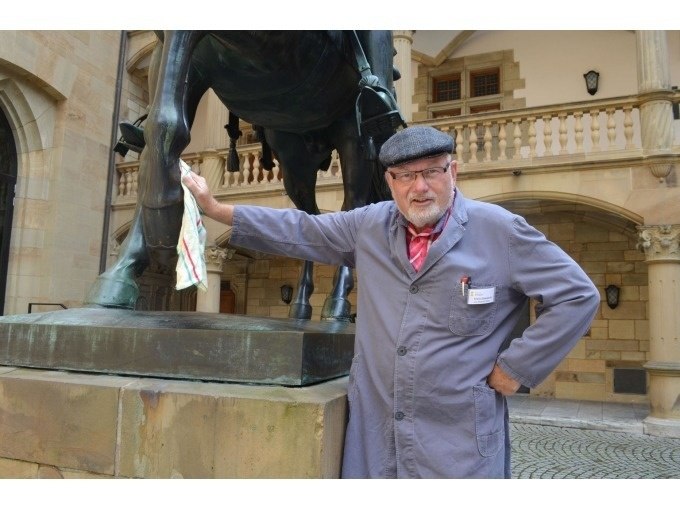 Ein Mann in blauer Arbeitskleidung und Mütze steht neben einer Pferdestatue und hält ein Tuch. Er befindet sich in einem historischen Innenhof., © Stuttgart Marketing GmbH
