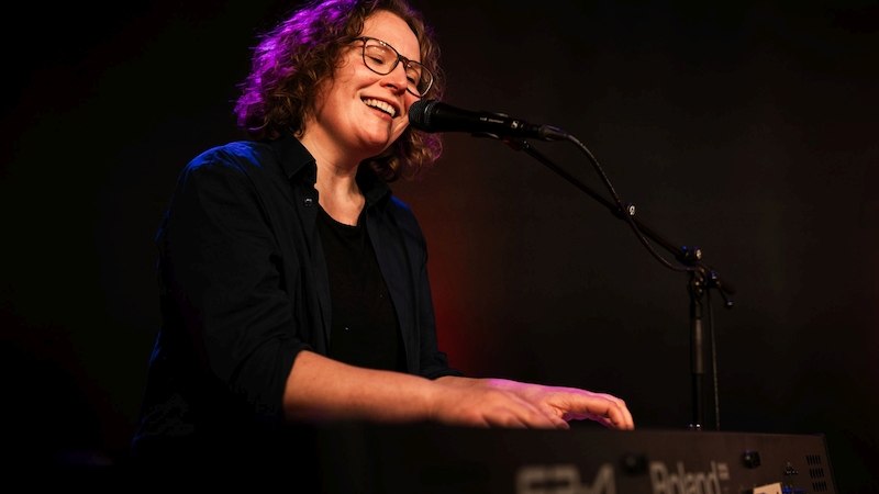 Eine Person mit Brille spielt lächelnd Keyboard und singt in ein Mikrofon auf einer Bühne., © Kulturhaus Schwanen