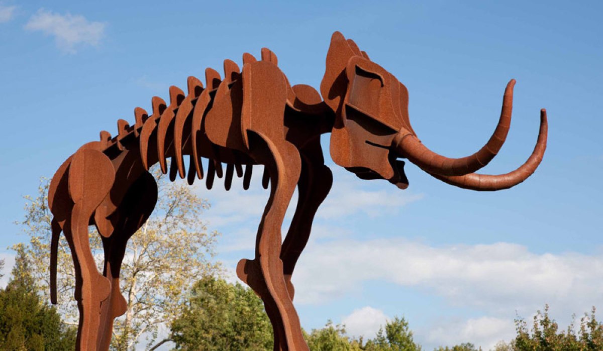 Lebensgro&szlig;e Mammutskulptur aus rostfarbenem Metall im Freien, umgeben von B&auml;umen und blauem Himmel., &copy; Hohenlohe + Schw&auml;bisch Hall Tourismus e.V.