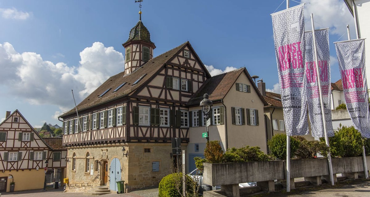 Das Alte Rathaus Waldenbuch mit Fachwerk und Türmchen, umgeben von Fahnen und blühenden Sträuchern, unter einem blauen Himmel mit Wolken., © Region Stuttgart Das Alte Rathaus Waldenbuch mit Fachwerk und Türmchen, umgeben von Fahnen und blühenden Sträuchern, unter einem blauen Himmel mit Wolken., © Region Stuttgart