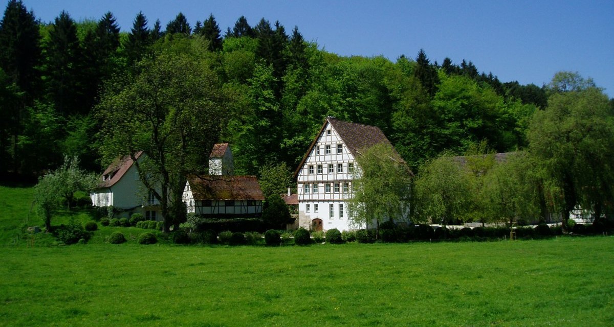 Fachwerkhäuser stehen in einer grünen Wiese vor einem dichten Wald. Der Himmel ist klar und blau., © Natur.Nah. Schönbuch & Heckengäu Fachwerkhäuser stehen in einer grünen Wiese vor einem dichten Wald. Der Himmel ist klar und blau., © Natur.Nah. Schönbuch & Heckengäu