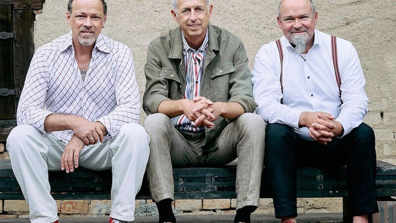 Drei M&auml;nner sitzen auf einer Bank vor einer rustikalen Wand. Sie tragen bunte Schuhe und l&auml;ssige Kleidung, die eine entspannte Atmosph&auml;re vermittelt., &copy; Mauerwerk GmbH