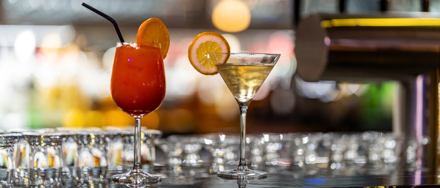 Zwei Cocktails auf einer Bar, einer rot mit Orangenscheibe, der andere klar mit Zitronenscheibe. Im Hintergrund unscharfe Lichter., © Plaza Residence Stuttgart-Ditzingen Zwei Cocktails auf einer Bar, einer rot mit Orangenscheibe, der andere klar mit Zitronenscheibe. Im Hintergrund unscharfe Lichter., © Plaza Residence Stuttgart-Ditzingen