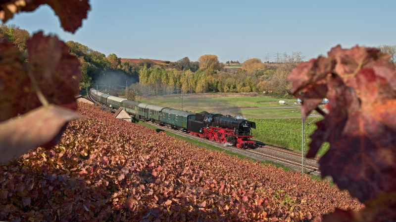 Eine Dampflok zieht einen Zug durch eine herbstliche Landschaft mit bunten Weinbergen und Bäumen., © DBK Historische Bahn e.V. Eine Dampflok zieht einen Zug durch eine herbstliche Landschaft mit bunten Weinbergen und Bäumen., © DBK Historische Bahn e.V.