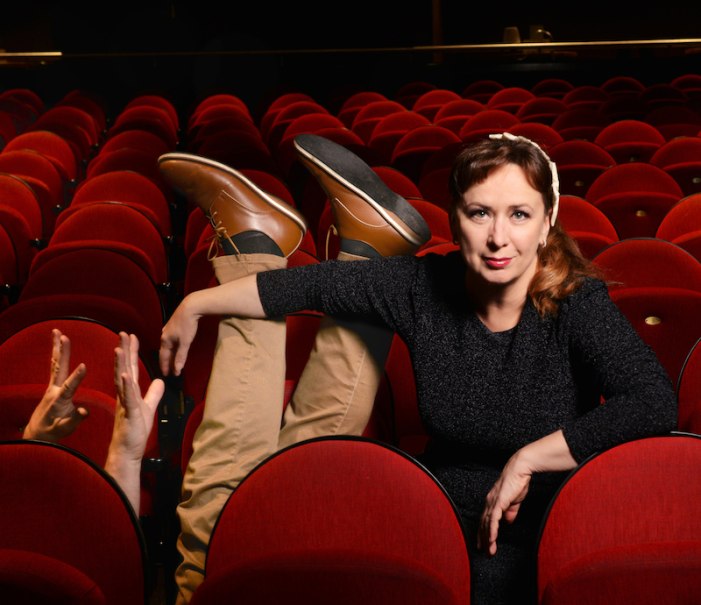 Frau sitzt in einem Theatersaal, während Beine und Hände einer anderen Person aus den roten Sitzen ragen. Humorvolle Szene., © Mauerwerk GmbH