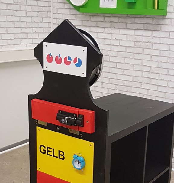 Escape-Room-Puzzle mit farbigen Schubladen und Symbolen. An der Wand eine grüne Dekoration mit Uhr und Rätseln., © Flucht-Raum