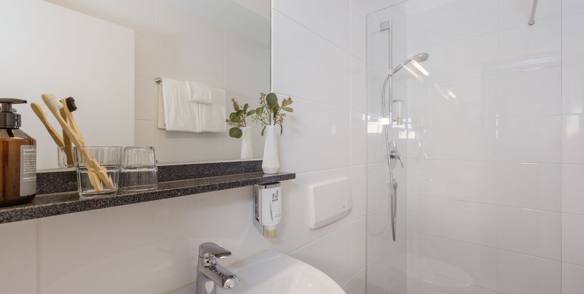 Modernes Badezimmer mit weiß gefliesten Wänden, Waschbecken, Spiegel, Glasregal mit Zahnbürsten und Duschbereich., © Novum Hospitality