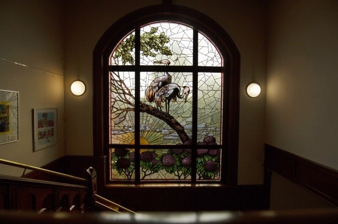 Ein kunstvolles Buntglasfenster mit Kranichen und Sonnenaufgang in einem historischen Treppenhaus, beleuchtet von zwei Wandlampen., &copy; Stadt G&ouml;ppingen