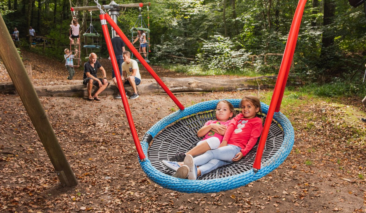 Zwei Kinder entspannen auf einer großen Netzschaukel im Waldspielplatz. Im Hintergrund spielen weitere Kinder, während Erwachsene auf einem Baumstamm sitzen. Zwei Kinder entspannen auf einer großen Netzschaukel im Waldspielplatz. Im Hintergrund spielen weitere Kinder, während Erwachsene auf einem Baumstamm sitzen.