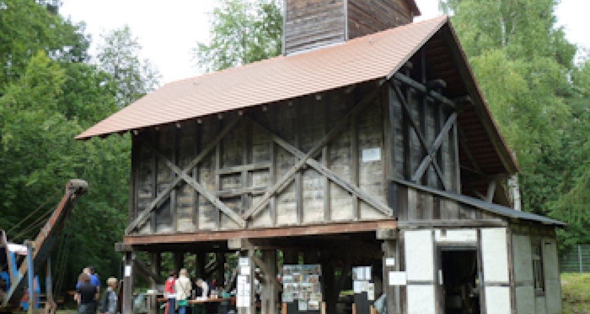 Historisches Sandwerk mit Holzkonstruktion und rotem Dach, umgeben von Bäumen. Menschen stehen davor und betrachten Ausstellungsstücke.