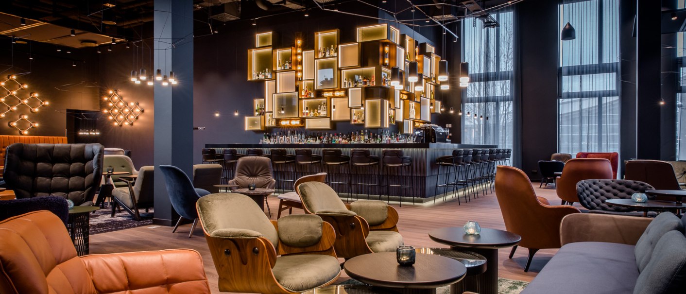 Elegante Bar mit modernen Möbeln, beleuchteten Regalen und gemütlicher Atmosphäre. Dunkle Farben und stilvolle Beleuchtung dominieren den Raum., © TOMAS