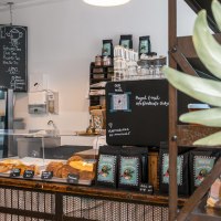 Ein gemütliches Café mit Theke, Gebäckauslage und Getränkekarte. Pflanzen schmücken den Raum, und eine Tafel zeigt das Menü an., © SMG, Sarah Schmid