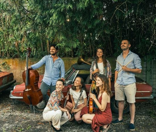 Sechs Musiker mit Instrumenten posieren vor einem alten Auto im Freien. Sie wirken fr&ouml;hlich und entspannt, umgeben von B&auml;umen., &copy; Foto_(c)_David Fichtner