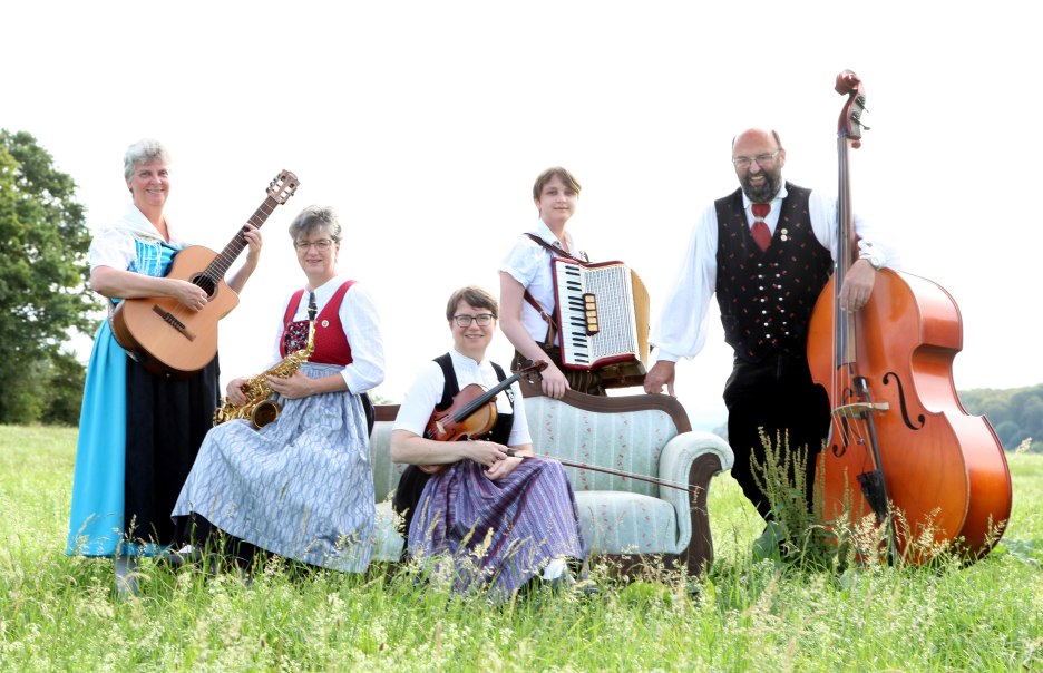 Eine Musikgruppe in traditioneller Kleidung posiert mit Instrumenten auf einer Wiese. Sie halten Gitarre, Saxophon, Geige, Akkordeon und Kontrabass.