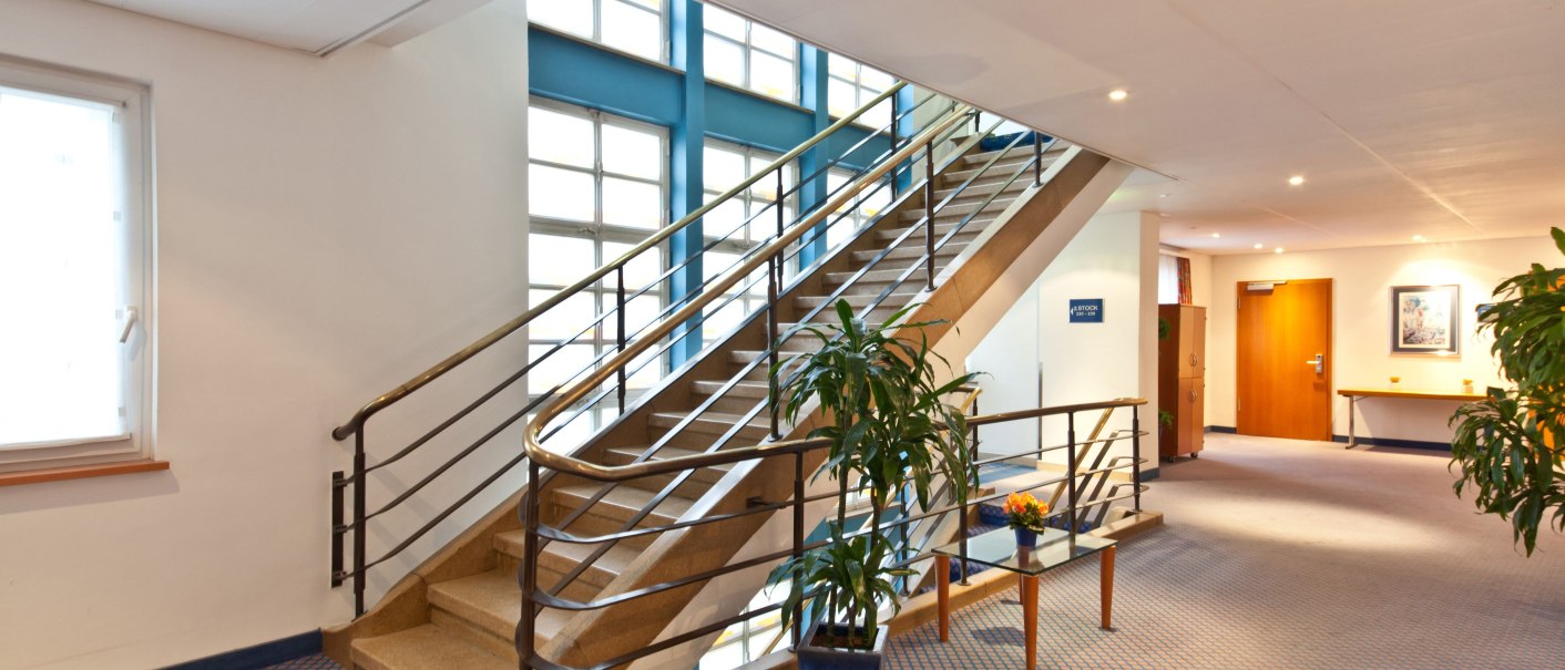 Ein modernes Treppenhaus mit Metallgeländer, Pflanzen in Töpfen und Teppichboden. Große Fenster lassen viel Licht herein., © Novum Boulevard Stuttgart City