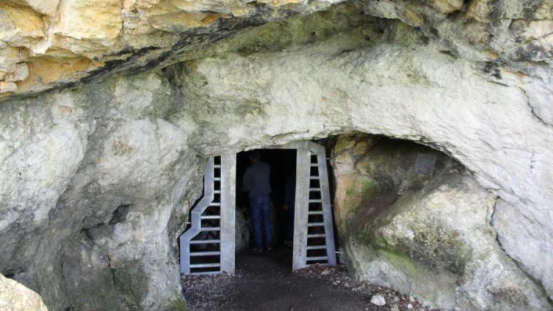 Ein H&ouml;hleneingang mit Metallgitter, durch den eine Person in die Dunkelheit geht. Die H&ouml;hle ist von Felsen umgeben.