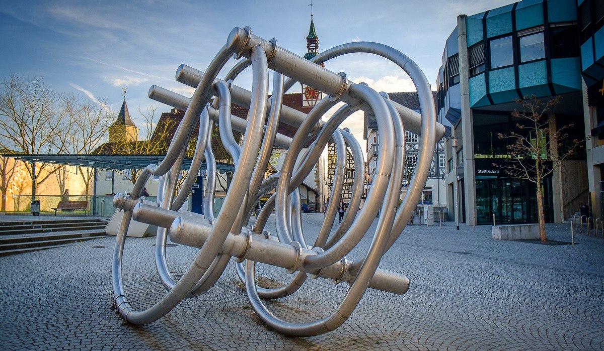 Metallskulptur aus verschlungenen Rohren auf einem Platz, umgeben von modernen und historischen Gebäuden., © Remstal Tourismus e.V.