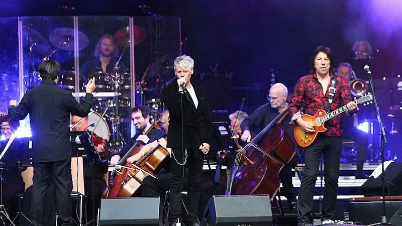 Ein S&auml;nger und ein Gitarrist performen mit einem Orchester auf einer B&uuml;hne. Der Dirigent leitet das Ensemble, w&auml;hrend die Musiker konzentriert spielen., &copy; Stadthalle Leonberg