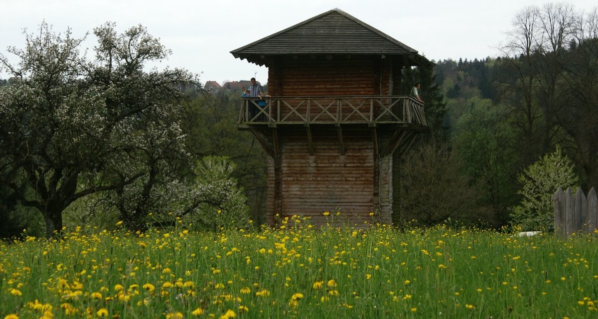 Ein hölzerner Limesturm auf einer Wiese mit gelben Blumen, umgeben von blühenden Bäumen und Wald., © Remstal Tourismus e.V. Ein hölzerner Limesturm auf einer Wiese mit gelben Blumen, umgeben von blühenden Bäumen und Wald., © Remstal Tourismus e.V.