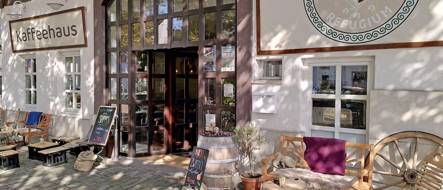 Gem&uuml;tliches Caf&eacute; mit Holzb&auml;nken und rustikaler Dekoration vor dem Eingang. Schilder und Pflanzen schaffen eine einladende Atmosph&auml;re., &copy; cKaffeehausMaLe