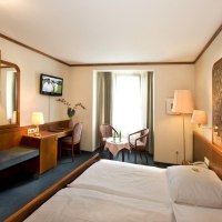 Gemütliches Hotelzimmer mit Doppelbett, Fernseher, Schreibtisch und Sitzbereich vor einem Fenster. Holzmöbel und warme Beleuchtung dominieren den Raum., © Hotel am Schelztor